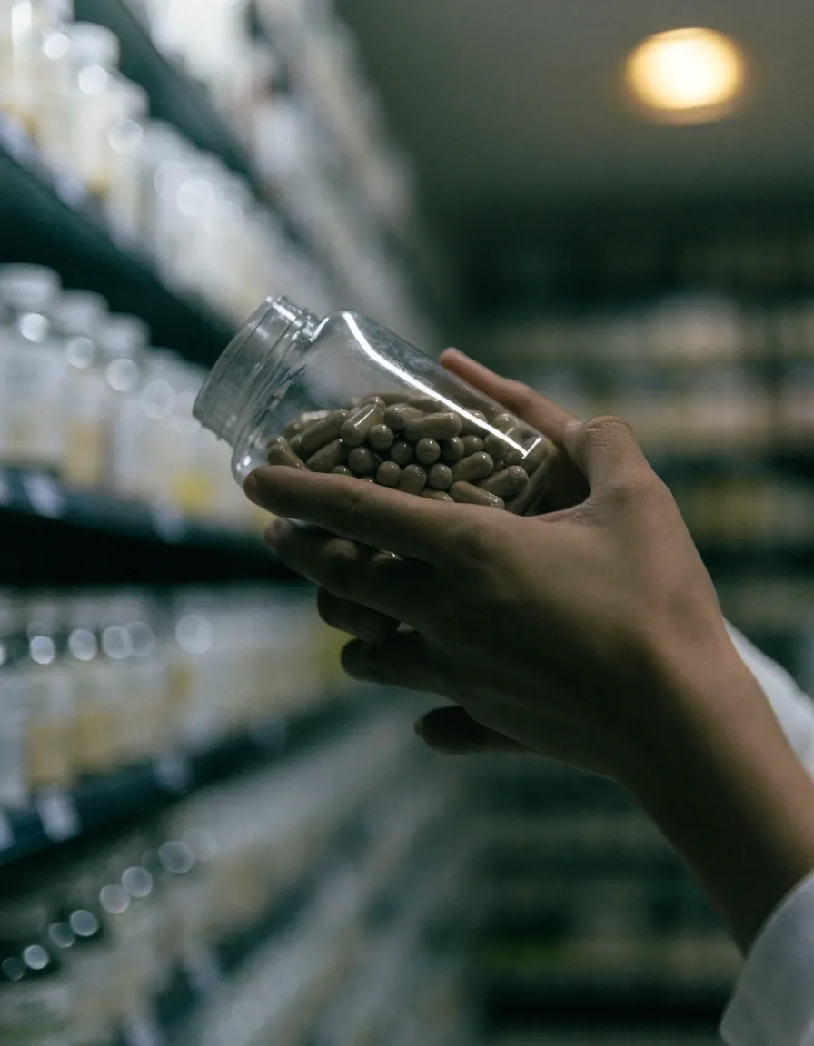 Hands turning an unmarked supplement bottle in a softly lit pharmacy aisle